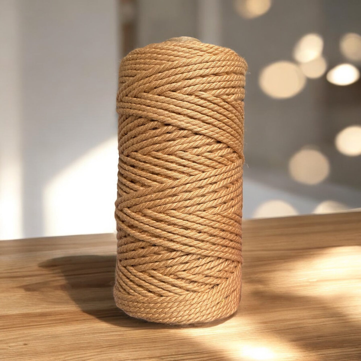 A spool of camel-coloured, ultra-soft 3mm 3ply Luxe Macrame Cotton stands upright on a wooden surface. The sunlight creates soft, circular bokeh effects in the blurred background, adding warmth to the scene.