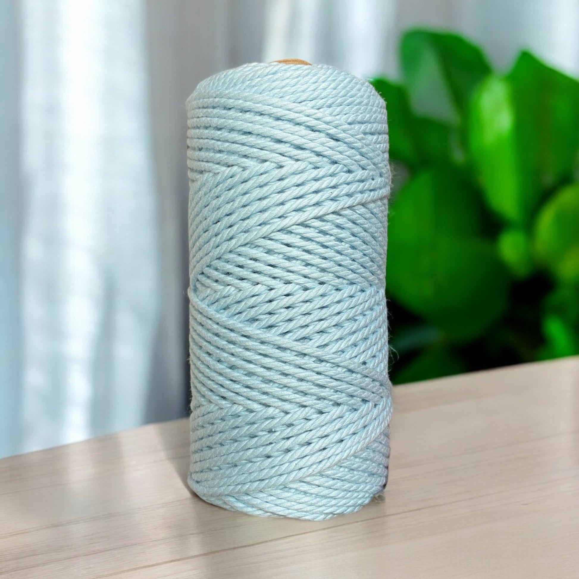 A spool of luxe premium cord in an ice blue colour is placed on a wooden table, with soft light casting shadows. In the background, green leaves are partially visible, adding a touch of nature to the scene.