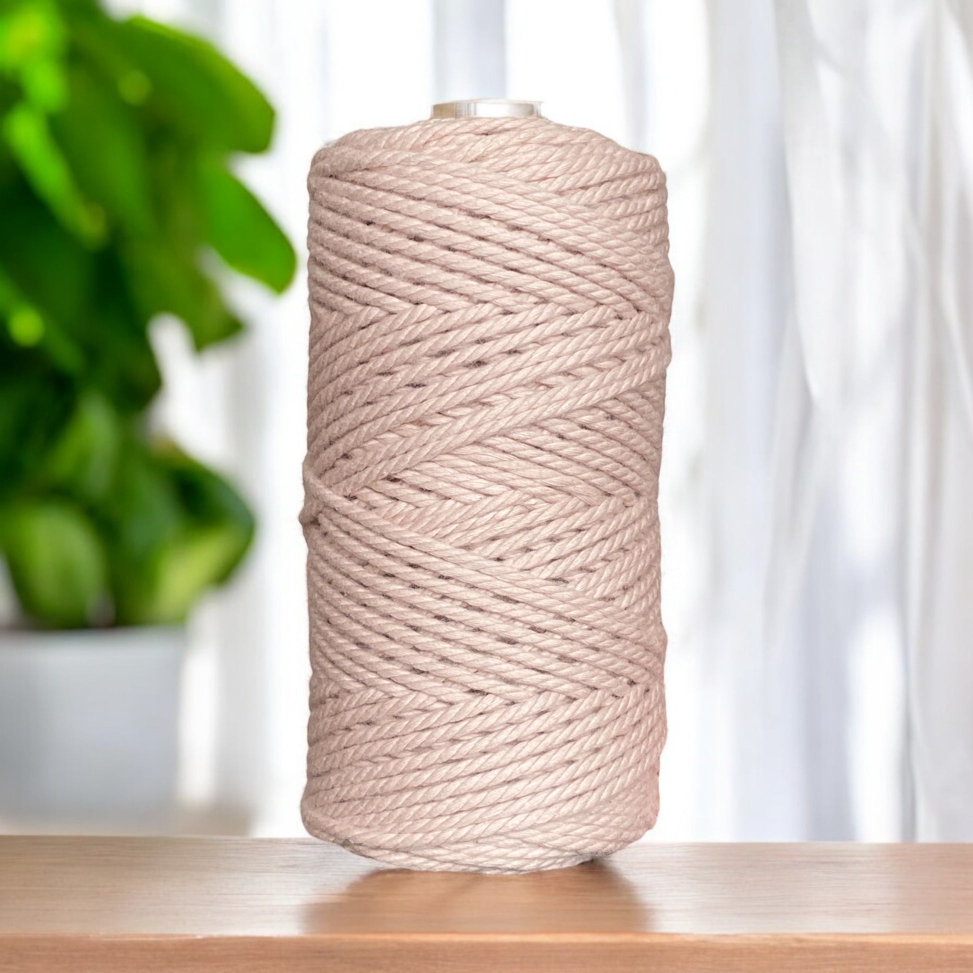 A large spool of 3mm 3ply macrame cord in a misty rose stands upright on a wooden surface. In the background, a plant with green leaves is visible near a softly lit white curtain, adding to the scene's inviting ambiance.