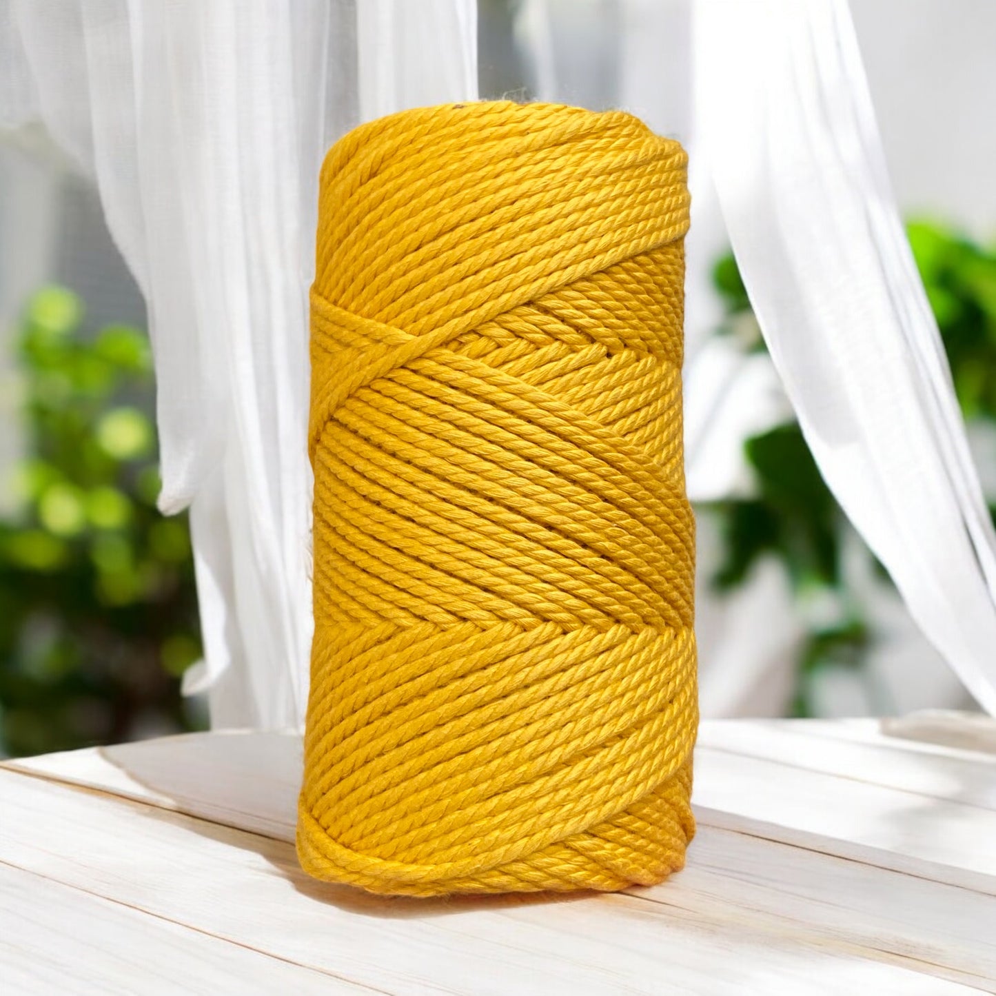 A large spool of bright yellow macrame cord sits on a wooden surface. White curtains and green foliage blur in the background, with sunlight creating a warm, inviting atmosphere.