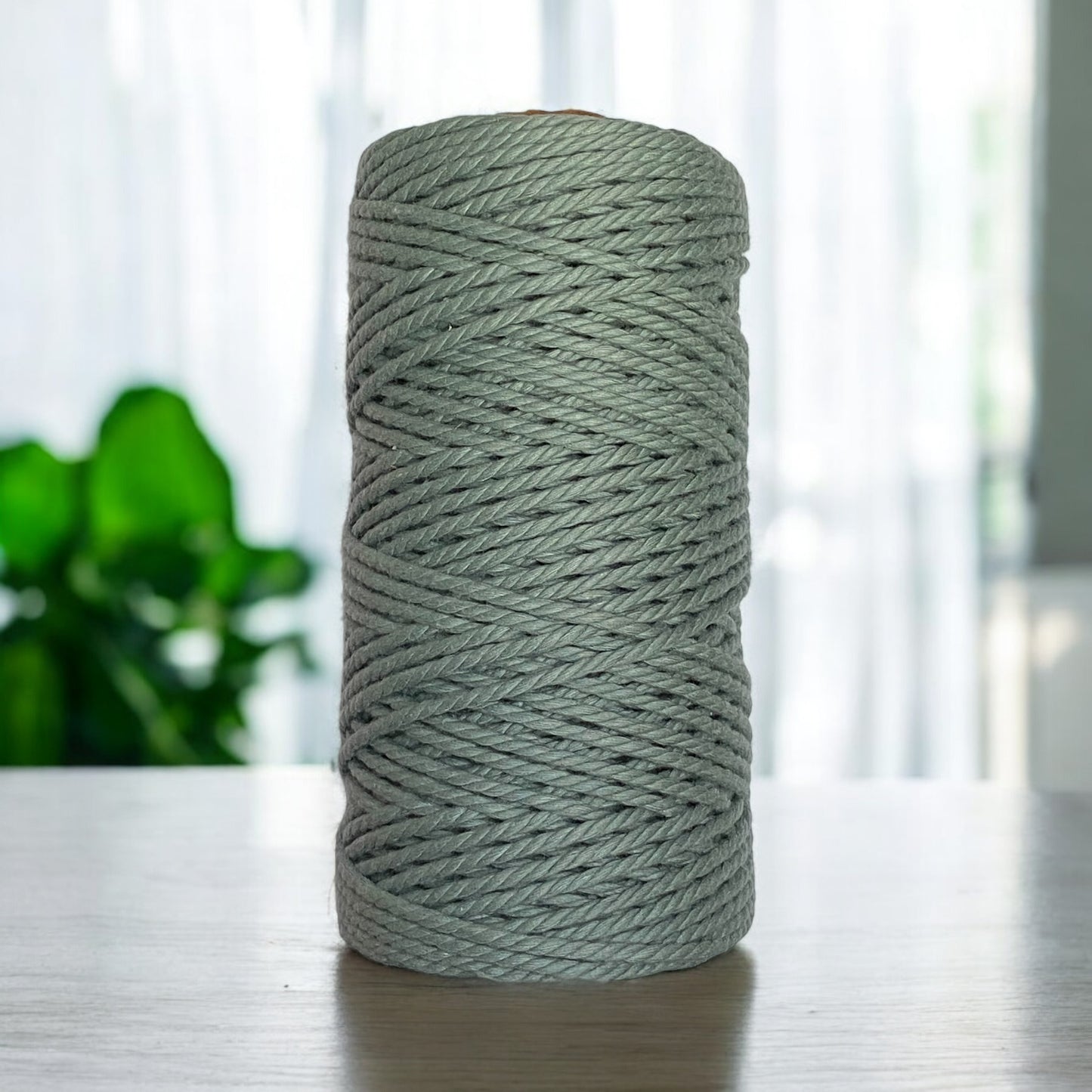 A spool of thick Cadet Blue 3mm 3ply Premium Luxe cord rests on a wooden table. The background reveals a blurred view of a window adorned with sheer curtains and a green potted plant, adding an elegant touch to the scene.