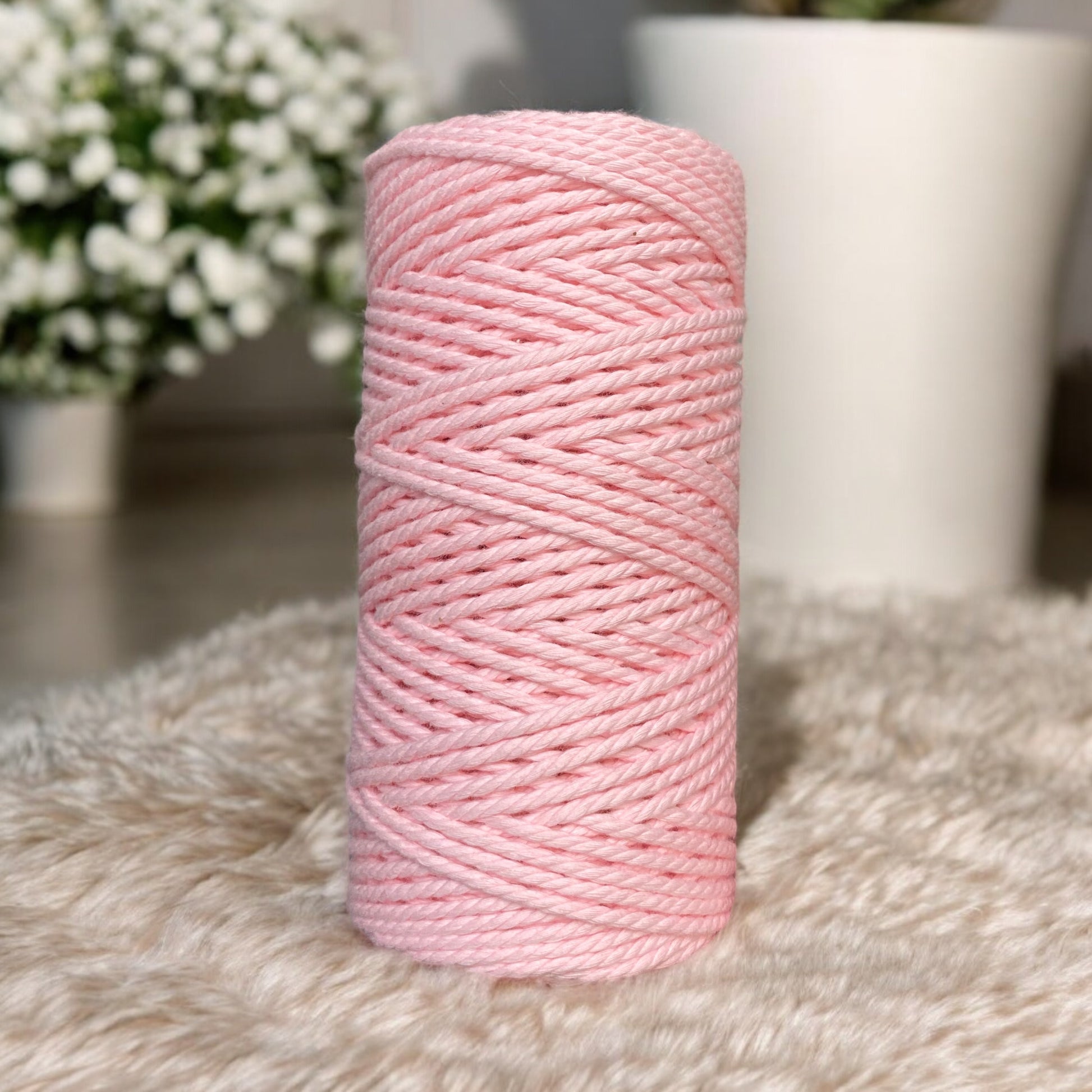 A spool of pink yarn stands upright on a soft, beige textured surface. In the blurred background, there are white flowering plants in pots. The scene conveys a cozy and serene atmosphere.