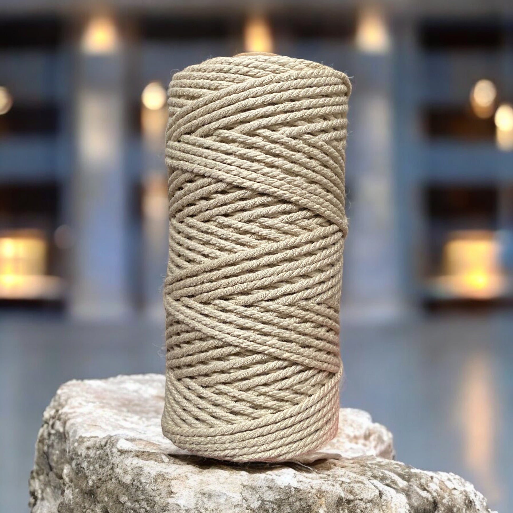 A spool of wheat-coloured, 3mm 3ply macrame cord stands upright on a textured stone surface. The background is softly blurred, with warm lights creating a cozy atmosphere.